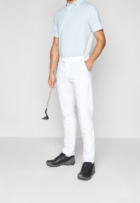 Světle modrá polo košile s krátkým rukávem a kruhovým vzorem, v kombinaci s bílými kalhotami a černými golfovými botami, držící golfovou hůl.