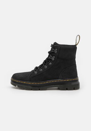 Zwarte suède veterlaars met dikke rubberen zool, gele stiksels, metalen oogjes en trekbandje aan de hiel.