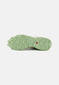 Semelle de chaussure de trail running avec une surface en caoutchouc texturé de couleur verte, arborant des crampons proéminents pour l'adhérence et un détail d'accent rouge.