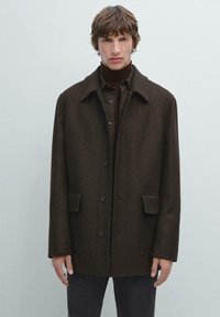 Cappotto in misto lana marrone con colletto, bottoni frontali e due tasche laterali. Tessuto testurizzato con vestibilità dritta, indossato sopra una camicia scura.