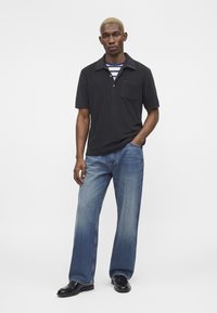 Homme aux cheveux blonds courts portant un polo noir sur un tee-shirt à rayures, jean bleu ample et chaussures noires, debout avec une main dans la poche.