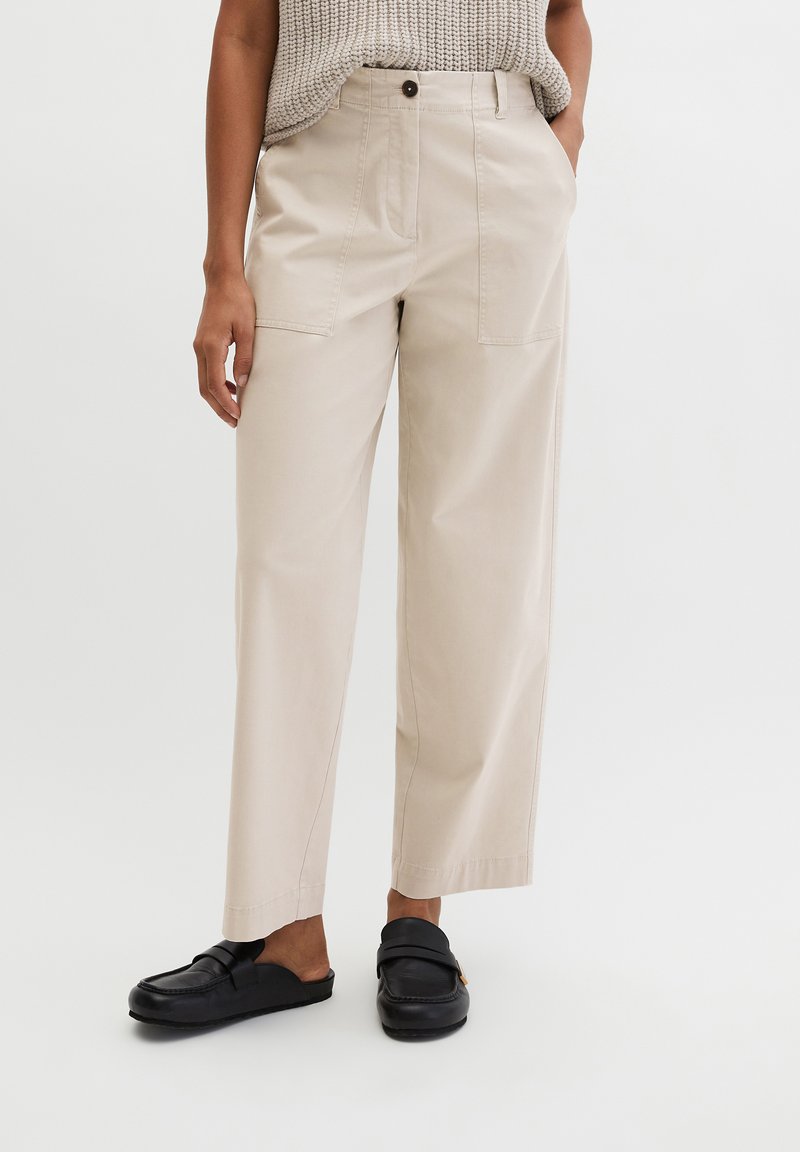 Γυναίκα που φοράει μπεζ παντελόνι με φαρδιά πόδια και εμπρόσθιες τσέπες, πλεκτό μπεζ τοπ χωρίς μανίκια και μαύρα δερμάτινα loafers χωρίς κορδόνια.