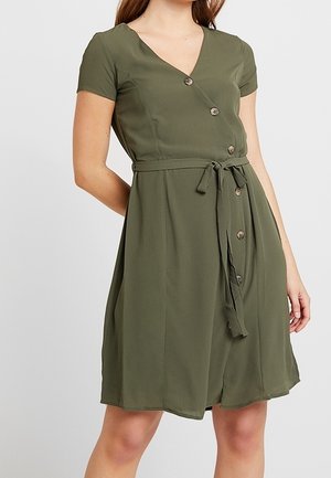 Kobieta w oliwkowej, krótkorękawowej sukience kopertowej sięgającej do kolan, z guzikami i wiązanym paskiem w talii.