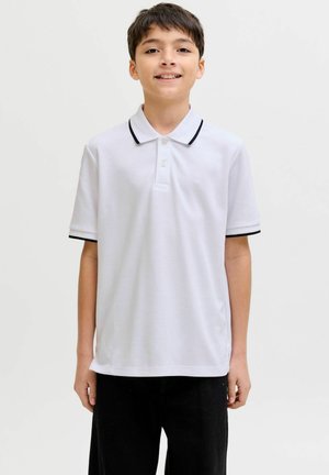 Usmievavý chlapec v bielom polokošeli s čiernym lemom a čiernych nohaviciach stojaci pred jednoduchým svetlým pozadím.