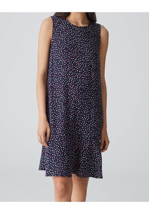 Robe bleu marine sans manches avec un motif de points roses et blancs dispersés, portée par une personne debout devant un fond gris clair uni.