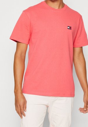 Jasnoróżowa, bawełniana koszulka z okrągłym dekoltem, krótkimi rękawami i małą naszywką z logo w górnym lewym rogu. Gładka tekstura.