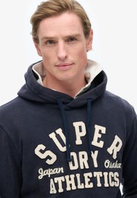 Granatowy hoodie z białym nadrukiem tekstu "SUPER DRY Japan Osaka ATHLETICS" w pogrubionej czcionce; posiada kaptur ze sznurkiem oraz warstwowy kołnierz.