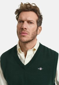 Groene gebreide vest met een V-hals ontwerp, gedragen over een witte blouse met kraag. Inclusief een klein geborduurd logo op de linkerkant van de borst.