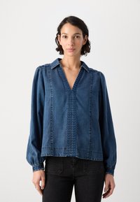 Denimbluse med V-hals, puffete lange ermer, og intrikate sømmer foran. Dyp blå farge, myk tekstur, og avslappet passform.