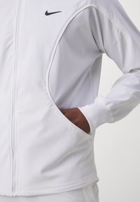 Weiße Sportjacke mit Reißverschluss, ausgestattet mit einem schwarzen Nike-Logo, seitlichen Taschen und gerippten Bündchen für einen sicheren Sitz und Komfort.