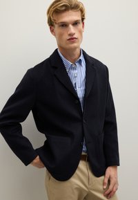 Giovane uomo che indossa un blazer nero sopra una camicia a righe blu e bianca e pantaloni beige, in piedi con una mano in tasca contro un muro semplice.