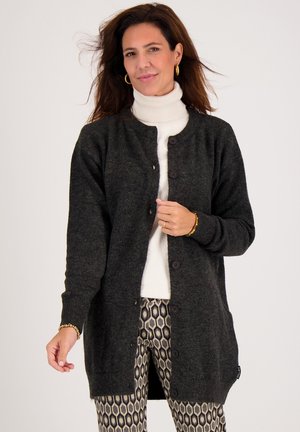 Donkergrijze gebreide cardigan met een ronde hals, knopen aan de voorkant en geribbelde manchetten. Draagt over een witte coltrui, gecombineerd met geblokte broeken.