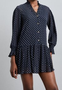 Mörkblå långärmad miniklänning med vita prickar, knappframsida, volangkrage och veckad kjoldel som bärs av en person.