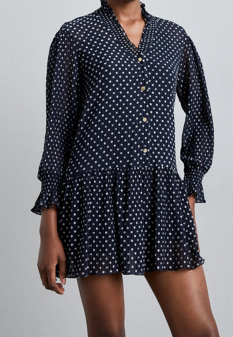 Mörkblå långärmad miniklänning med vita prickar, knappframsida, volangkrage och veckad kjoldel som bärs av en person.