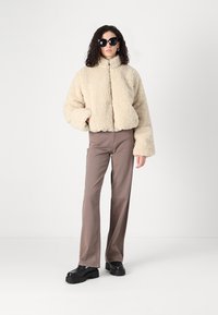 Beige faux fur bunda s vysokým límcem a zipovým zapínáním, doplněná o volné hnědé kalhoty a černé platformové boty. Černé sluneční brýle dotvářejí celkový vzhled.