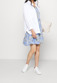 Biała koszula zapinana na guziki noszona na niebieskiej sukience w kwiatowy wzór, zestawiona z białymi trampkami i teksturowaną beżową kopertówką.