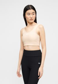 Νεαρή γυναίκα που φοράει μπεζ κοντό τοπ και μαύρο κολάν ψηλόμεσο, στέκεται γυρίζοντας το κεφάλι της προς τα αριστερά.