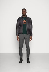 En sort puffersjakke med lynlås foran; en grøn t-shirt med orange grafisk tryk; grå skræddersyede bukser; sorte snørebåndsstøvler. Modellen står foran en hvid baggrund.