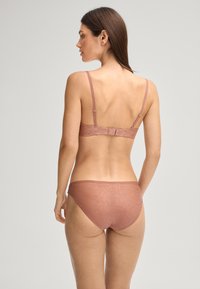 Nainen, joka käyttää matching mauve-väristä liivejä ja alushousuja, joissa on kukkakuvioita, näkyy takaa yksinkertaista taustaa vasten.