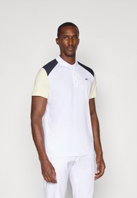 Biała koszulka polo z krótkimi rękawami, jasnoniebieskimi akcentami i granatowymi panelami na ramionach. Posiada klasyczny kołnierzyk oraz logo na piersi.