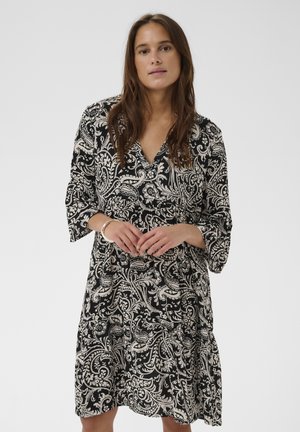 Žena s dlhými hnedými vlasmi, oblečená v čierno-bielych šatách s paisley vzorom po kolená a s tromištvrťovými rukávmi, stojaca pred jednoduchým pozadím.