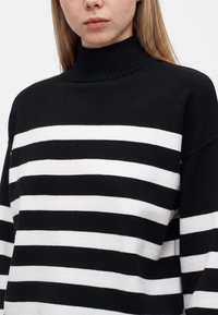 Femme portant un pull à col roulé à rayures horizontales noires et blanches, montrée des épaules au menton sur un fond uni.