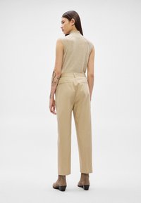 Γυναίκα με μακριά σκουρόχρωμα μαλλιά και τατουάζ στα χέρια, ντυμένη με μπεζ αμάνικο τοπ, ψηλόμεσα καφέ παντελόνια και καφέ μποτάκια μέχρι τον αστράγαλο, στέκεται με την πλάτη.