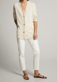 Frau trägt beige Strickjacke mit Knöpfen, weiße schmale Hose und schwarze flache Sandalen, steht mit einer Hand in der Tasche vor neutralem Hintergrund.