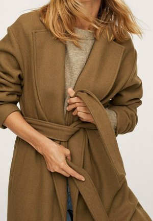 Žena si zavazuje pásek na hnědém vlněném kabátu přes béžový svetr, s rozházenými blond vlasy a zastrčenými rukávy.