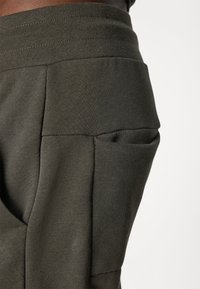 Lähikuva tummanoliivinvihreistä collegehousuista, joissa on leveä joustava vyötärönauha ja suuri sivutasku läpällä.