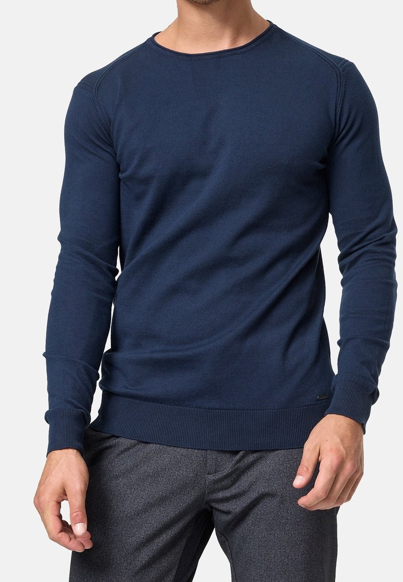 Homme portant un pull bleu marine à col rond et manches longues et un pantalon gris foncé, debout devant un fond clair uni.