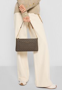 Brown patterned handbag with a zip closure, gold hardware, and a chain strap. The material appears textured, complementing cream-colored wide pants.