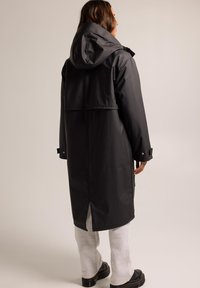 Manteau noir imperméable avec capuche, doté d'une large ventilation dans le dos, de poignets boutonnés et d'une conception de superposition à la taille.
