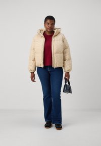 Giacca a vento imbottita crema con motivo a diamante e cappuccio, abbinata a una camicia bordeaux, jeans blue e scarpe nere. Piccola borsa nera in mano.
