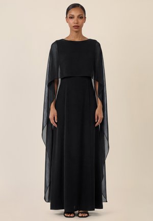 Frau, die nach vorne blickend steht, trägt ein langes schwarzes Kleid mit fließenden, durchsichtigen Cape-Ärmeln und schwarze Sandalen mit offenem Zehenbereich vor einem beigen Hintergrund.