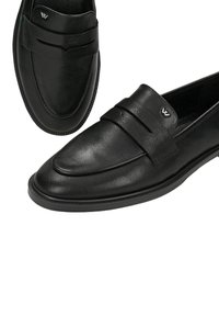 Μαύρα δερμάτινα μοκασίνια slip-on με λεπτομέρεια ιμάντα και ένα μικρό ασημί λογότυπο στο επάνω μπροστινό μέρος κάθε παπουτσιού.
