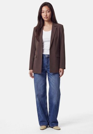 Femme portant un blazer marron à fines rayures, un haut blanc, un jean bleu à jambes larges et des chaussures beiges, debout devant un fond clair uni.