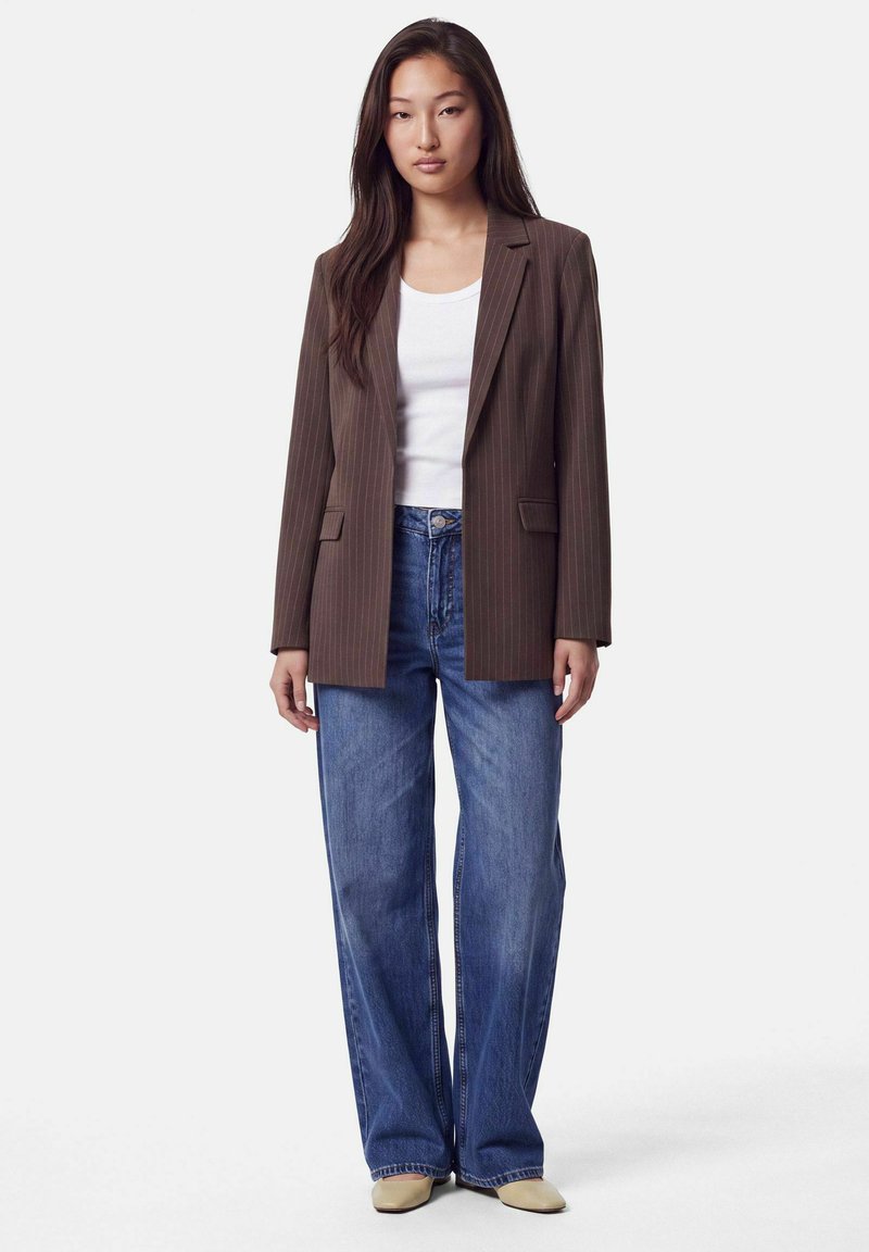 Femme portant un blazer marron à fines rayures, un haut blanc, un jean bleu à jambes larges et des chaussures beiges, debout devant un fond clair uni.