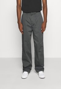 Grijze, losvallende broeken met een gladde textuur, voorzien van een ritssluiting en voorplooien, gecombineerd met witte sneakers. Zwart shirt zichtbaar.