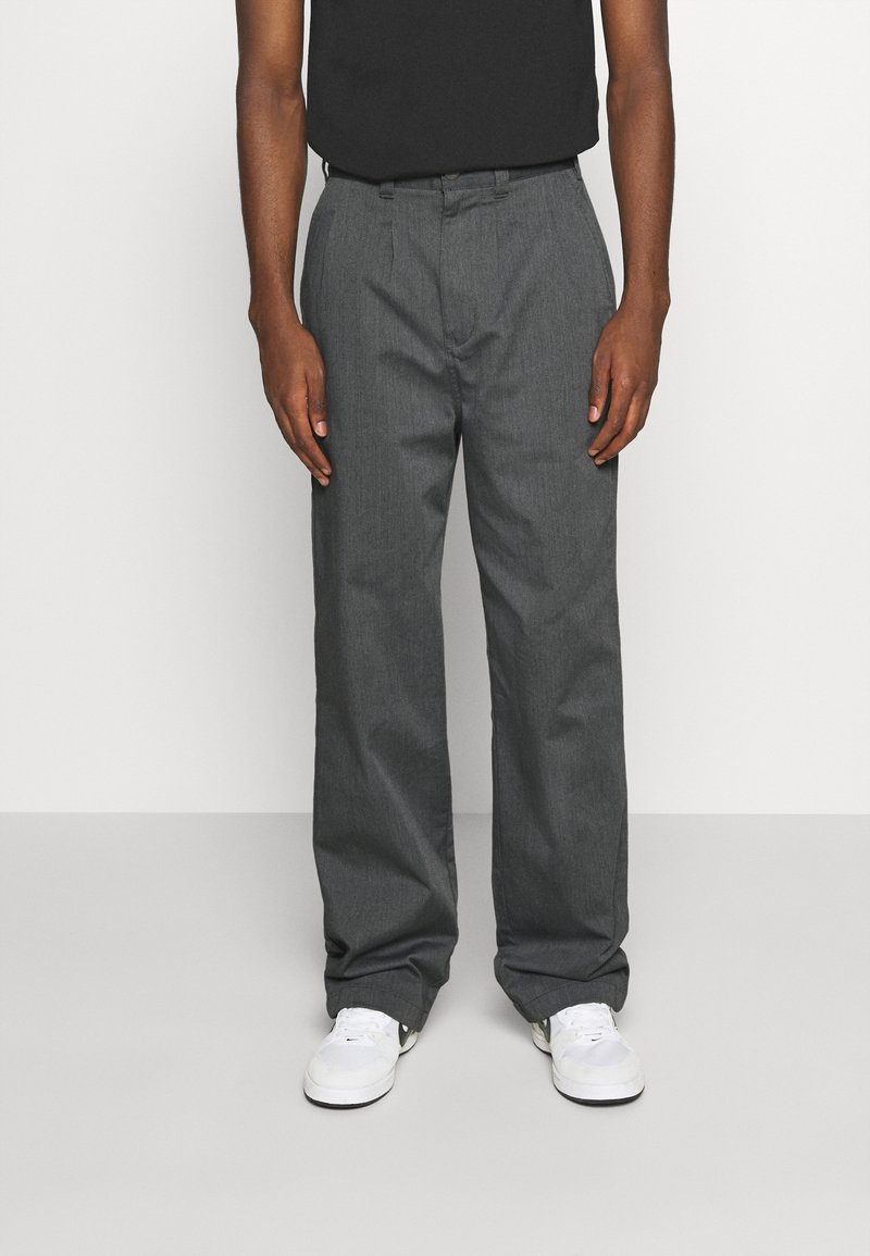 Grijze, losvallende broeken met een gladde textuur, voorzien van een ritssluiting en voorplooien, gecombineerd met witte sneakers. Zwart shirt zichtbaar.