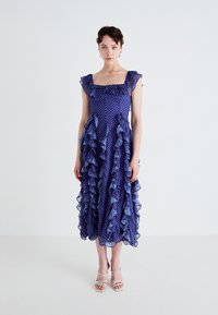 Un vestido arrugado, de longitud a la rodilla, en violeta con puntos blancos. Presenta un escote cuadrado y detalles de volantes transparentes en capas.