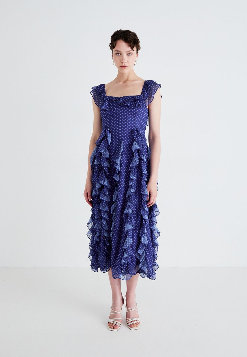 Un vestido arrugado, de longitud a la rodilla, en violeta con puntos blancos. Presenta un escote cuadrado y detalles de volantes transparentes en capas.