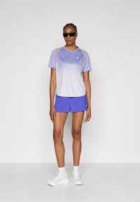 Αθλητική μπλούζα σε ανοιχτό μωβ χρώμα με σχεδιασμό gradient, συνδυασμένη με μπλε σορτς. Περιλαμβάνονται γυαλιά ηλίου και λευκά αθλητικά παπούτσια. Λείο ύφασμα, άνετη εφαρμογή.