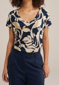 Marineblaues und cremefarbenes T-Shirt mit kurzen Ärmeln und floralem Muster mit Blättern; weicher Stoff mit einem entspannten V-Ausschnitt-Design. Kombiniert mit marineblauen Hosen.