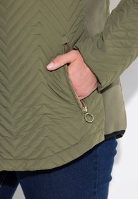 Olijfgroene jas met een doorgestikt chevronpatroon, voorzien van een ritszak en gladde stof, gecombineerd met donkerblauwe jeans.