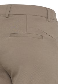Gros plan sur l'arrière d'un pantalon beige montrant une poche passepoilée, des passants de ceinture et des coutures visibles sur un tissu lisse.
