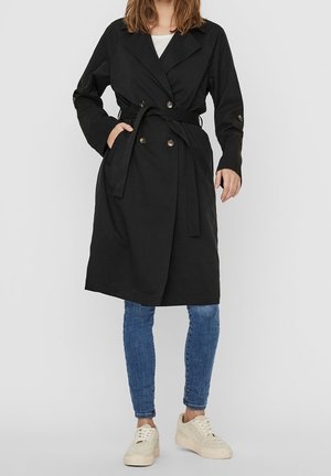 Femme portant un trench-coat noir ceinturé, un jean skinny bleu, des baskets blanches, debout devant un fond clair uni.
