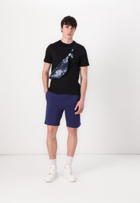 Fekete, rövid ujjú póló, sötétkék rövidnadrág, fehér sportcipő. Kék, tye-dye válltáska, állítható pánttal. Egyszerű dizájn, laza illeszkedés.