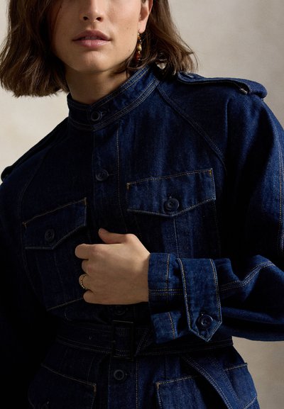 Ženska nosi temno modro jeans jakno z gumbki na prsnih žepih in pasom, drži jakno blizu prsi.