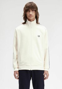 Chaqueta de chándal cremosa con cremallera, cuello alto, mangas raglán, bolsillos laterales y un pequeño logotipo en el pecho. Textura suave.
